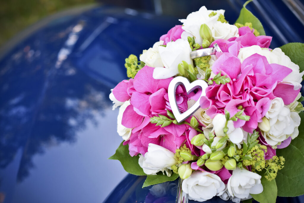 Brudbukett med rosa och vita blommor, med ett hjärta och vigselringarna placerade på motorhuven av en blå bil. Romantisk bröllopsbild med unika detaljer. Fotograf Martina Ardbäck