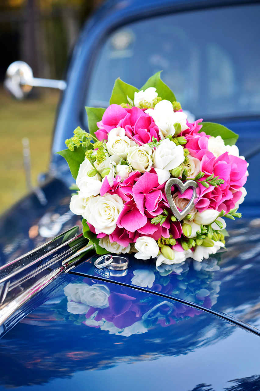 Brudbukett med rosa och vita blommor, med ett hjärta och vigselringarna placerade på motorhuven av en blå bil. Romantisk bröllopsbild med unika detaljer. Fotograf Martina Ardbäck